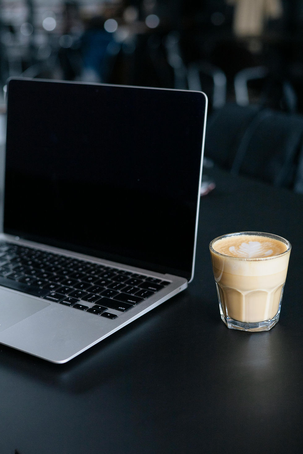 Laptop and latte with heart design on black table. Blurred background gives a calm café vibe. No visible text.