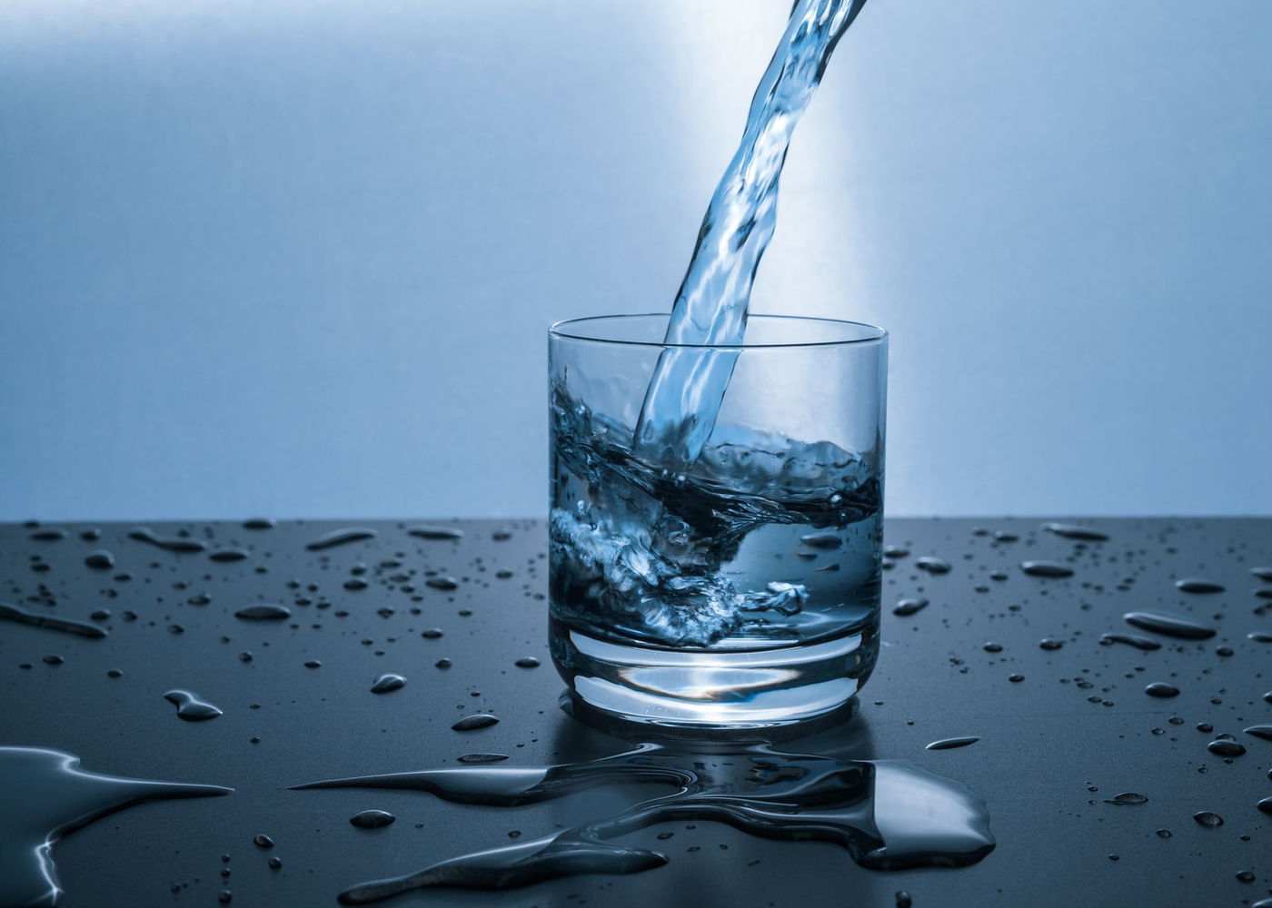 Clear water streams into a clear short tumbler on a wet reflective surface, blue studio background. No people, branding, product or home depicted.