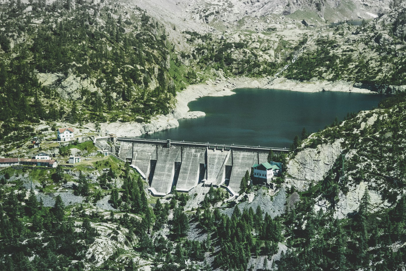 Large concrete dam wall, dark reservoir, rocky alpine hills and conifers, small utility buildings.