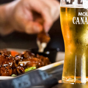 The image features a plate of food accompanied by glasses of Molson Canadian beer. The setting appears to be indoors, likely at a bar or restaurant, where food and drink are being enjoyed together. The scene captures the essence of dining and socializing with alcohol in a casual atmosphere.