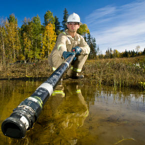 A man is standing in a river, wearing a white hard hat and holding a fishing pole. The scene is set outdoors with a clear sky above and trees nearby. The water reflects the surrounding environment.