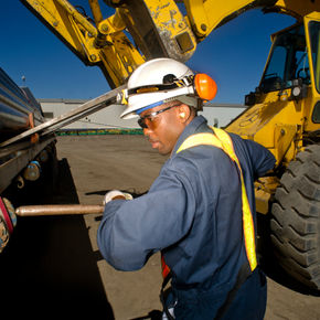 A man is wearing a hard hat and safety goggles while holding a piece of wood. He appears to be outdoors, likely at a construction site, surrounded by various construction equipment. The scene suggests he is engaged in blue-collar work, indicative of an engineering or construction role.
