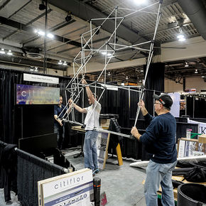 Men are seen working in a factory setting. The environment is indoor, featuring elements like computers and engineering tools. The individuals are dressed casually in jeans and footwear, indicating a practical work attire suitable for the factory environment.