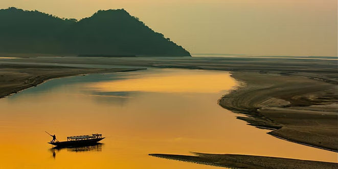 A small boat crossing the golden Brahmaputra River at sunset beneath dark hills in Assam, India.