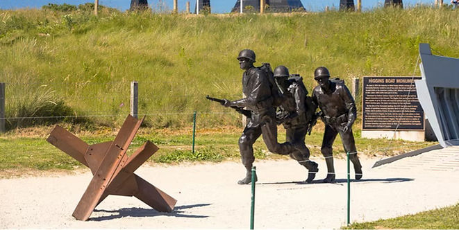 Bronze figures of advancing soldiers passing a beach obstacle at a Normandy D-Day memorial in France.