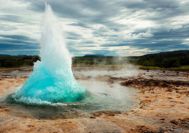 Geyser erupting from a geothermal basin with bright blue water and cloudy sky overhead.