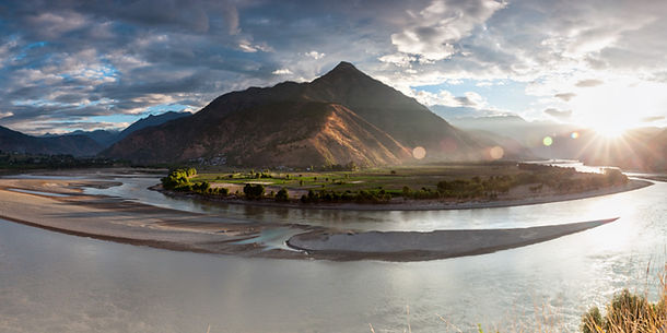 Mountain valley landscape with a winding river and sunlight breaking through dramatic clouds.