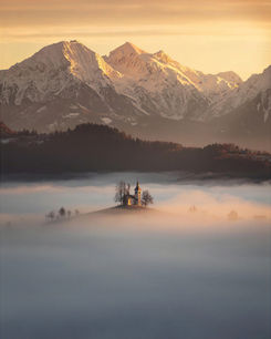 Small island in misty water beneath snow-capped mountains glowing in soft sunrise light.