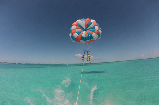 Two travelers parasailing beneath a colorful parachute over turquoise Caribbean water in Grand Turk.