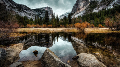 Mountain cliffs, evergreen forest, and autumn vegetation reflected in calm water.