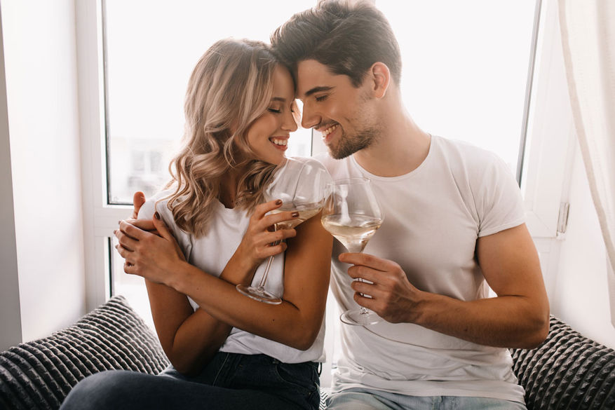Pareja feliz sonriendo, brindando con copas de vino cerca de la ventana invitaciones digitales.