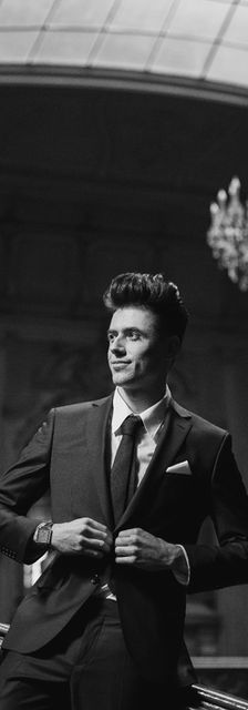 Elegant man in suit posing, with chandelier in background. Promocionales Alexa.