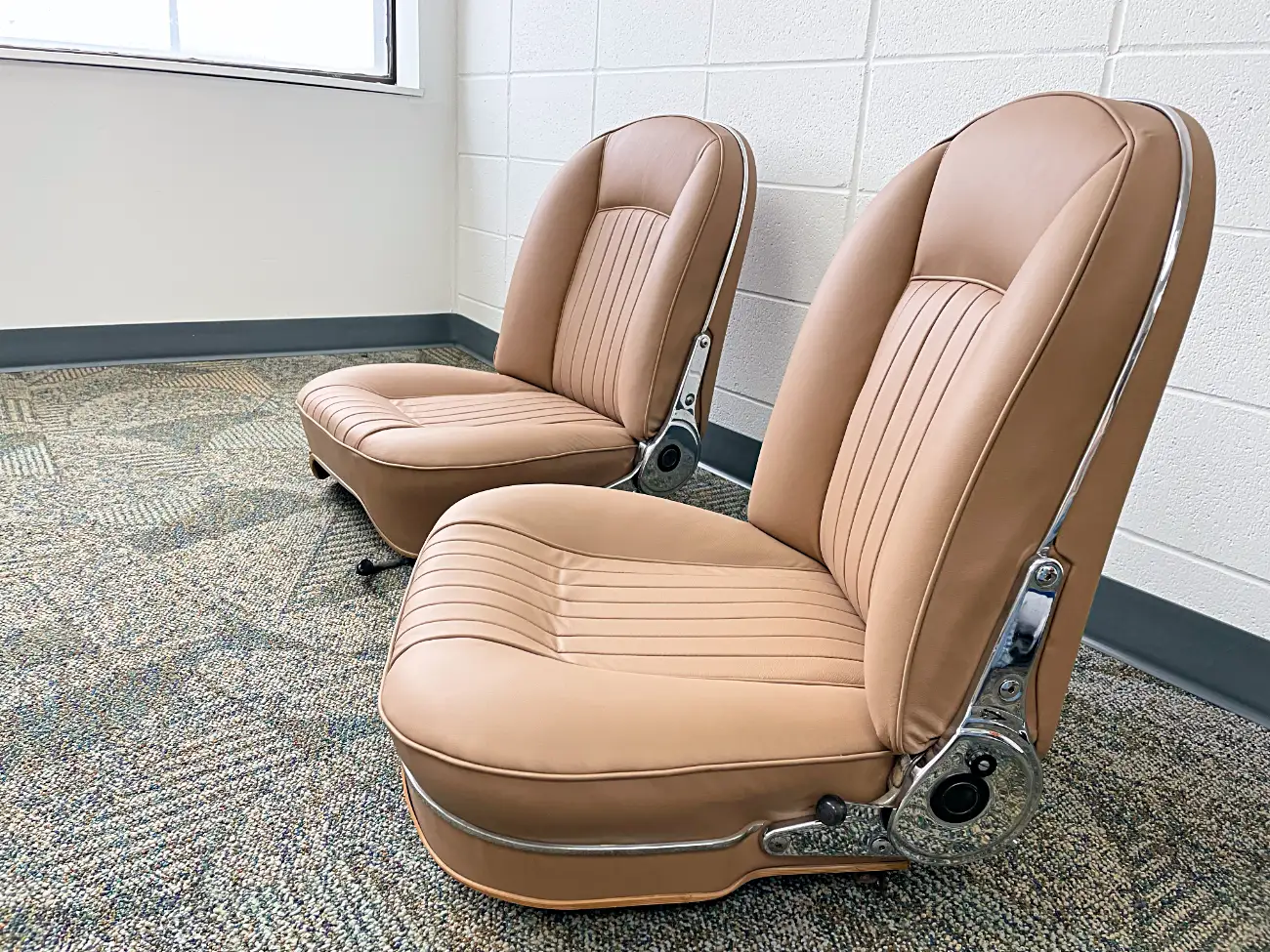 Two tan car seats on a carpeted floor in a room with white brick walls and a window, creating a vintage and nostalgic vibe.
