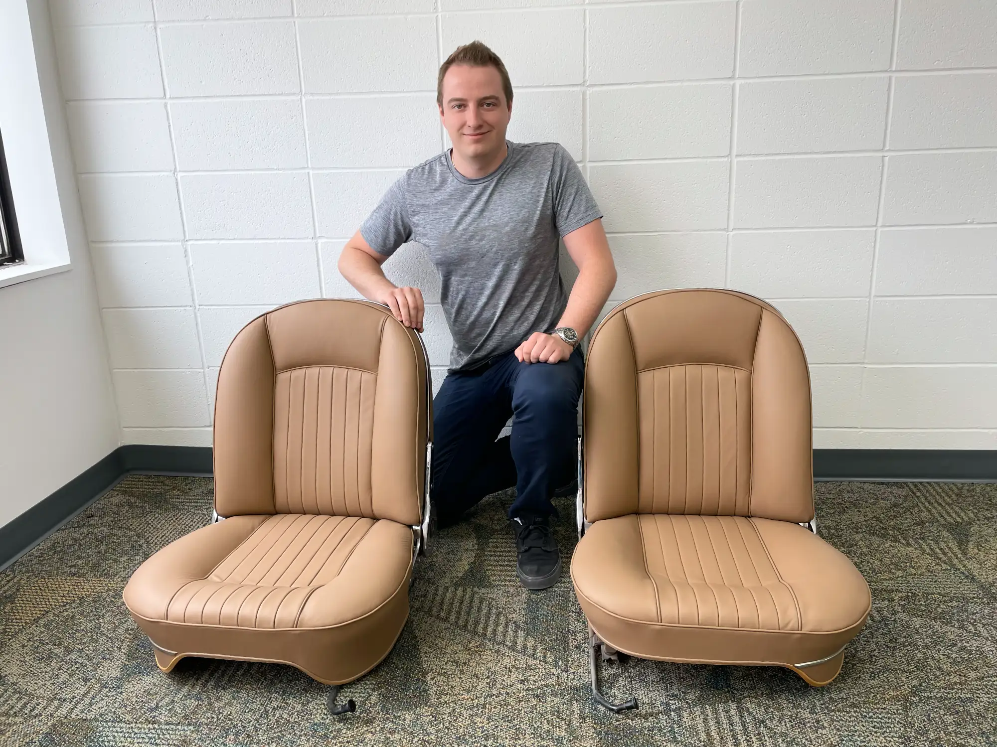 Young man kneeling by two tan car seats in an office with white brick walls and gray carpet. He smiles, suggesting satisfaction or pride.
