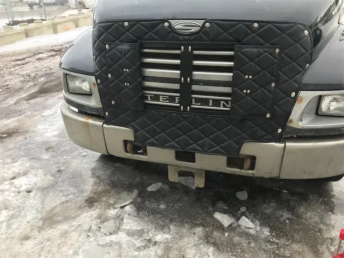 Front of a Sterling truck with a black quilted grille cover parked on icy, snowy pavement.