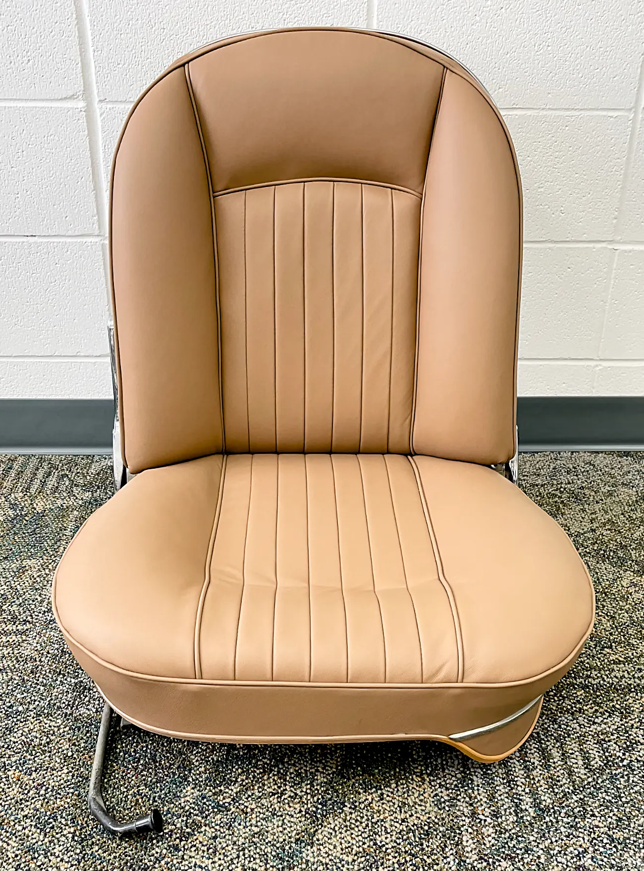 Tan leather chair with vertical stitching on a speckled carpet. White tiled wall background. Simple and retro design.