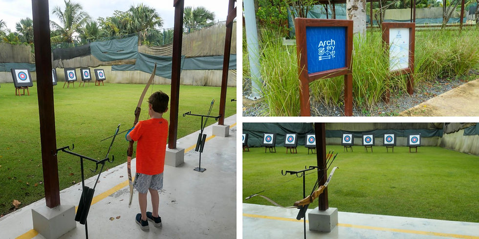 Boy in orange shirt practices archery outdoors, aiming at targets on green grass. Nearby sign reads "Archery." Lush trees in background.