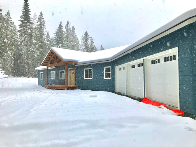 The front of a blue house with three garage door and some windows in the front as well as an overbuild on the entrance surrounded by snow in Sandpoint Idaho.