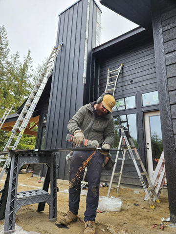 Construction worker grinding metal to finish the siding and trim of a dark grey house in the woods in Sandpoint Idaho.