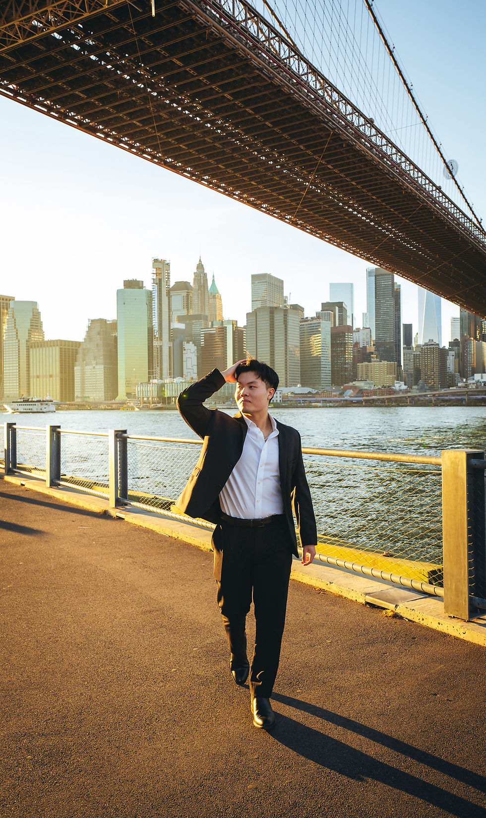 Dating app photographer client in New York City, Asian man in a suit walking under the Brooklyn Bridge with the Manhattan skyline behind him