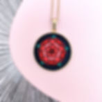 Collier représentant une rose rouge brodée sur du tissu noir en plus gros plan et avec un fond d'image rose et texturé.