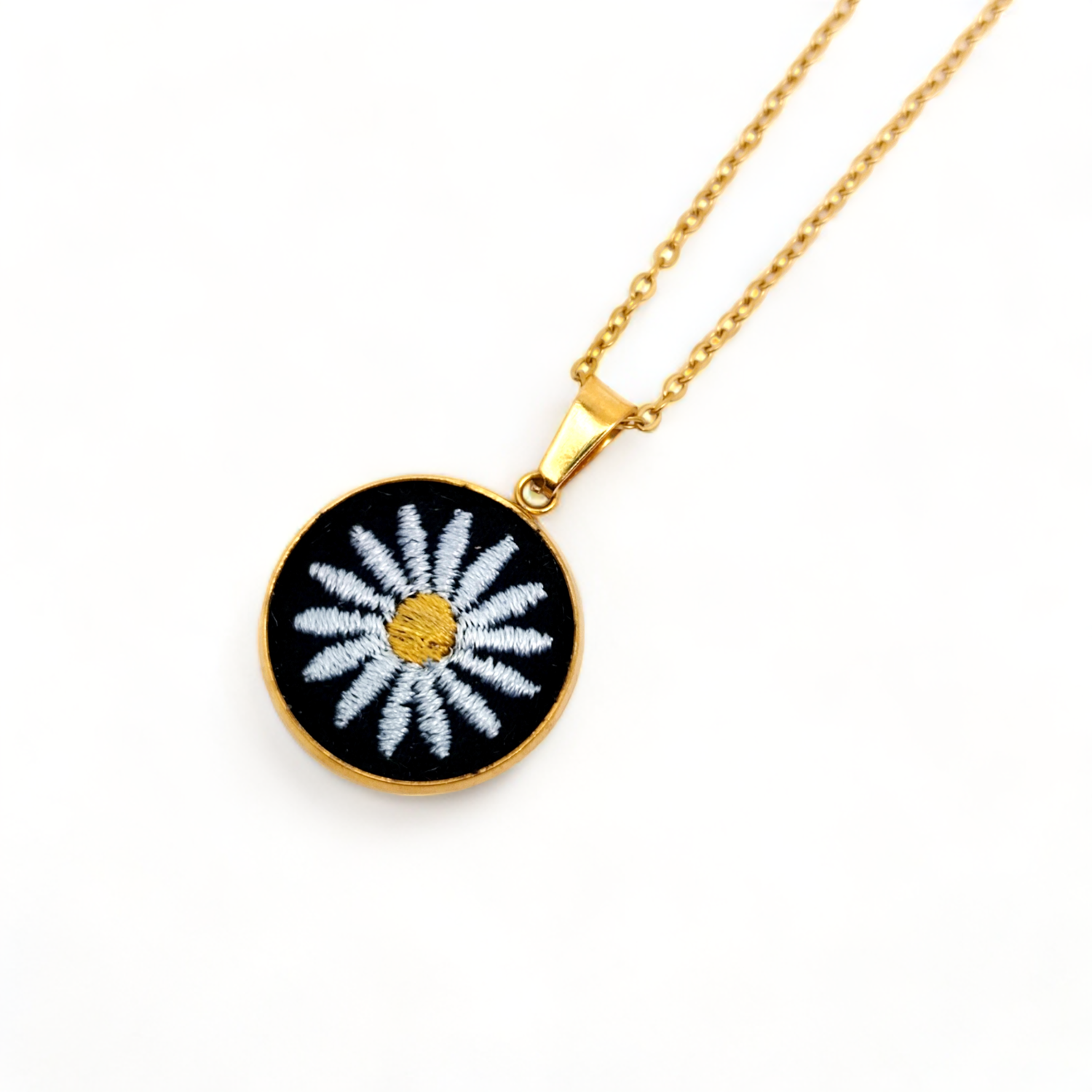 Photo du collier marguerite. On voit une marguerite blanche en broderie sur fond noir, sur un médaillon doré, avec la chaîne