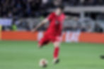 Soccer player in red kit with number 8 strikes a ball on a grassy field. Background has blurred audience and orange advertising boards.