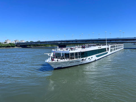 River cruise ship Scenic Amber sailing under a blue sky with passengers on the sun deck.