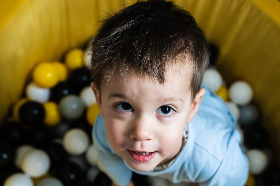 Un enfant souriant en pull bleu dans une piscine à balles jaune, noire et blanche. Fond jaune vif. Ambiance joyeuse.