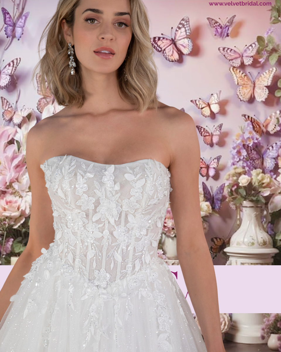 Bride wearing the strapless Tessa gown by Velvet Bridal, featuring a floral lace bodice with 3D appliqués and a full glitter-tulle ballgown skirt. The soft pink and lavender butterfly backdrop enhances the romantic, ethereal feel of the gown.
