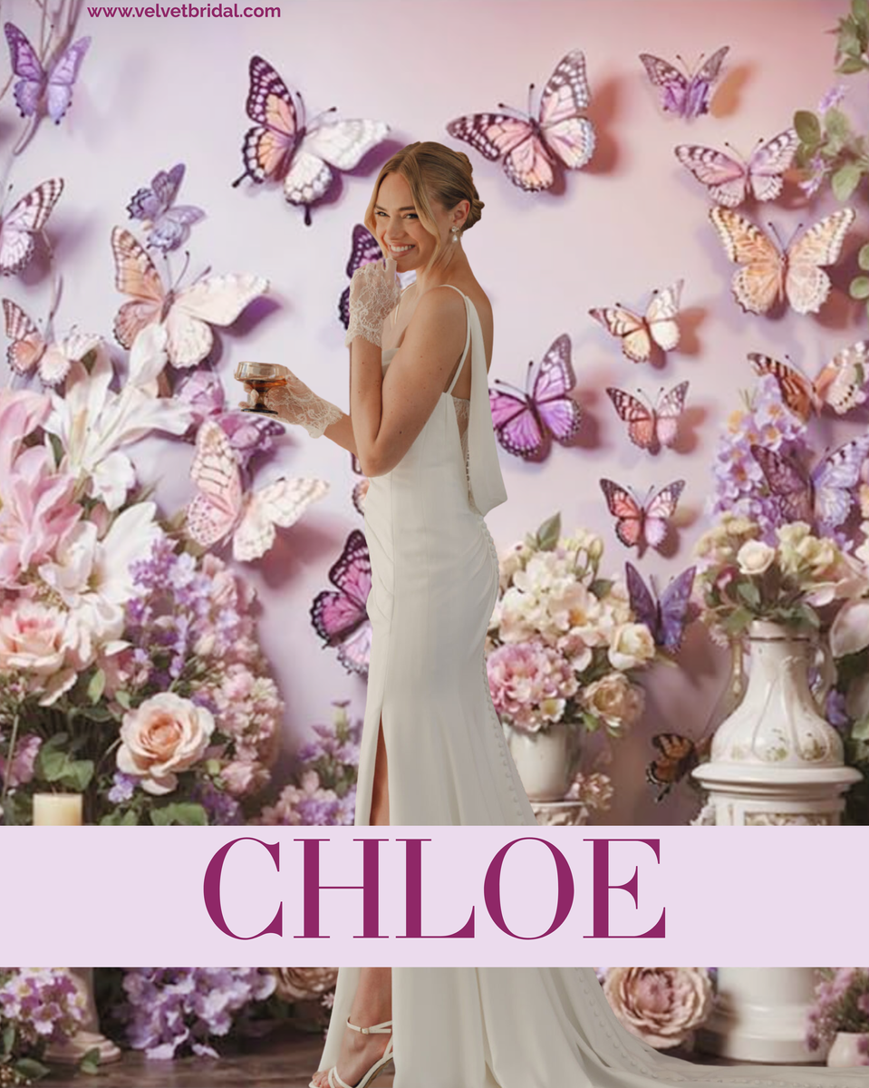 Bride wearing the Chloe wedding gown from Velvet Bridal (Stella York 8224) — a sleek crepe sheath with dainty spaghetti straps, a softly draped scoop neckline, asymmetrical ruching, a detachable cowl back with sheer lace, and a row of buttons trailing down the train. Styled in a soft, romantic floral butterfly backdrop.