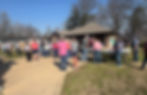Community gathering outdoors for an auction, people in casual clothes near tables with food. Bright day with clear skies and trees in the background.