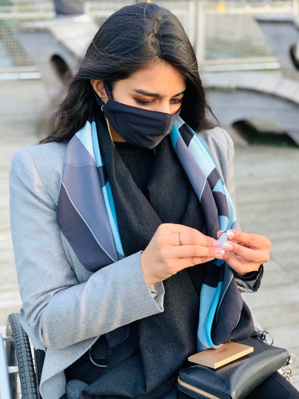 Sonia is a beautiful Indian women with long straight brown hair. Sonia is holding one of our dress weights which is a black circle weight with a sticker-like, adhesive backing. She is peeling the sticker off. She is wearing heather gray blazer, black sweater, and beautiful geometric pattern silk scarf with turquoise, gray, and taupe colors. She is also wearing a black face mask. She has her black wallet on her lap and the case for her dress weights on top. She is seated outside in her manual wheelchair.