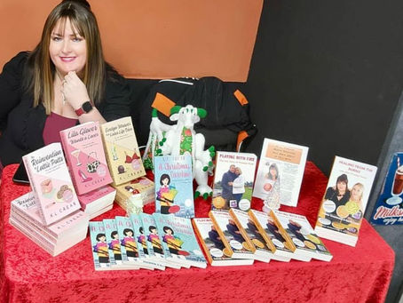 A woman sits at a table with various books displayed on a red cloth. The setting is indoors with an orange and black wall backdrop.