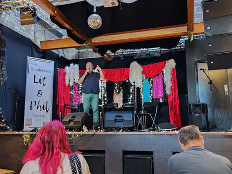 A speaker addresses the audience at a Sunderland Lit & Phil Society event, held in a charming, rustic venue adorned with vibrant backdrop and stage lights.