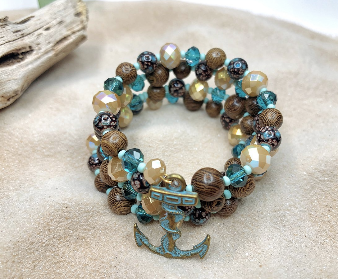 Handmade nautical memory wire bracelet with turquoise crystals and anchor charm on sand with driftwood background.