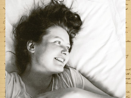 Close-up of a young woman lying on a pillow, smiling softly, looking off-camera. A handwritten signature in pink at the top edge.