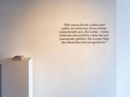 Gallery wall with a German-language quotation in large dark lettering. A white plinth in the foreground holds a copy of the book.