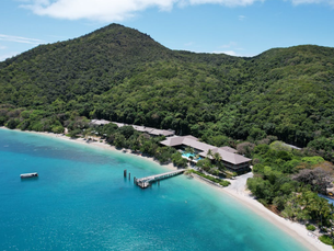 An aerial view of a tropical island shoreline with clear blue water, a sandy beach, a jetty extending into the sea, and resort buildings nestled at the base of lush green hills.