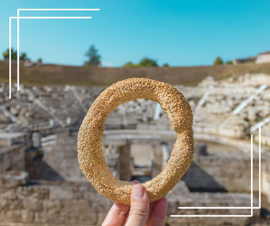 Hand holding a sesame koulouri against the backdrop of an ancient amphitheater under a clear blue sky, with modern white border lines.