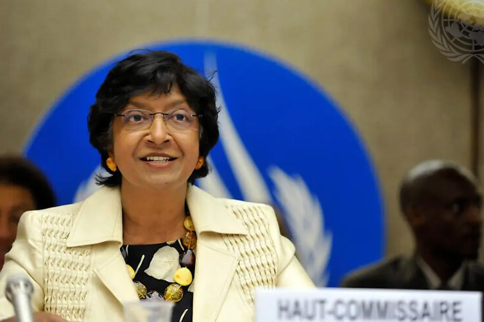 Navi Pillay wears a yellow jacket and stands in front of a blue and grey background