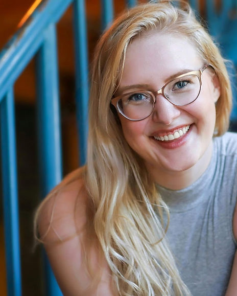 Madeline, a pale white woman, smiles at the camera while leaning slightly forward. She has wavy blonde hair and glasses and is wearing a grey sleeveless top. In the background is a blue hand railing.