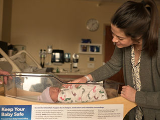 Woman leans over newborn in a hospital bassinet, smiling; a sign in front reads Keep Your Baby Safe.