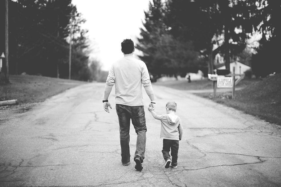 Black-and-white rear view of a man holding a young boy’s hand as they walk down a quiet tree-lined road.