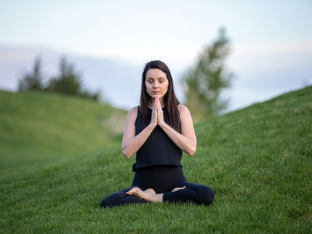 Woman in black yoga wear meditates cross-legged on green grass, eyes closed and hands in prayer under a soft sky.