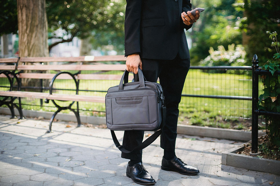 Man in a black suit holds a phone and gray briefcase in a sunny park. He stands near a wooden bench on a paved path. Lush greenery in background.