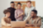 A team of five professionals working together, looking at one team member's laptop, with a cardboard whiteboard in the backgr