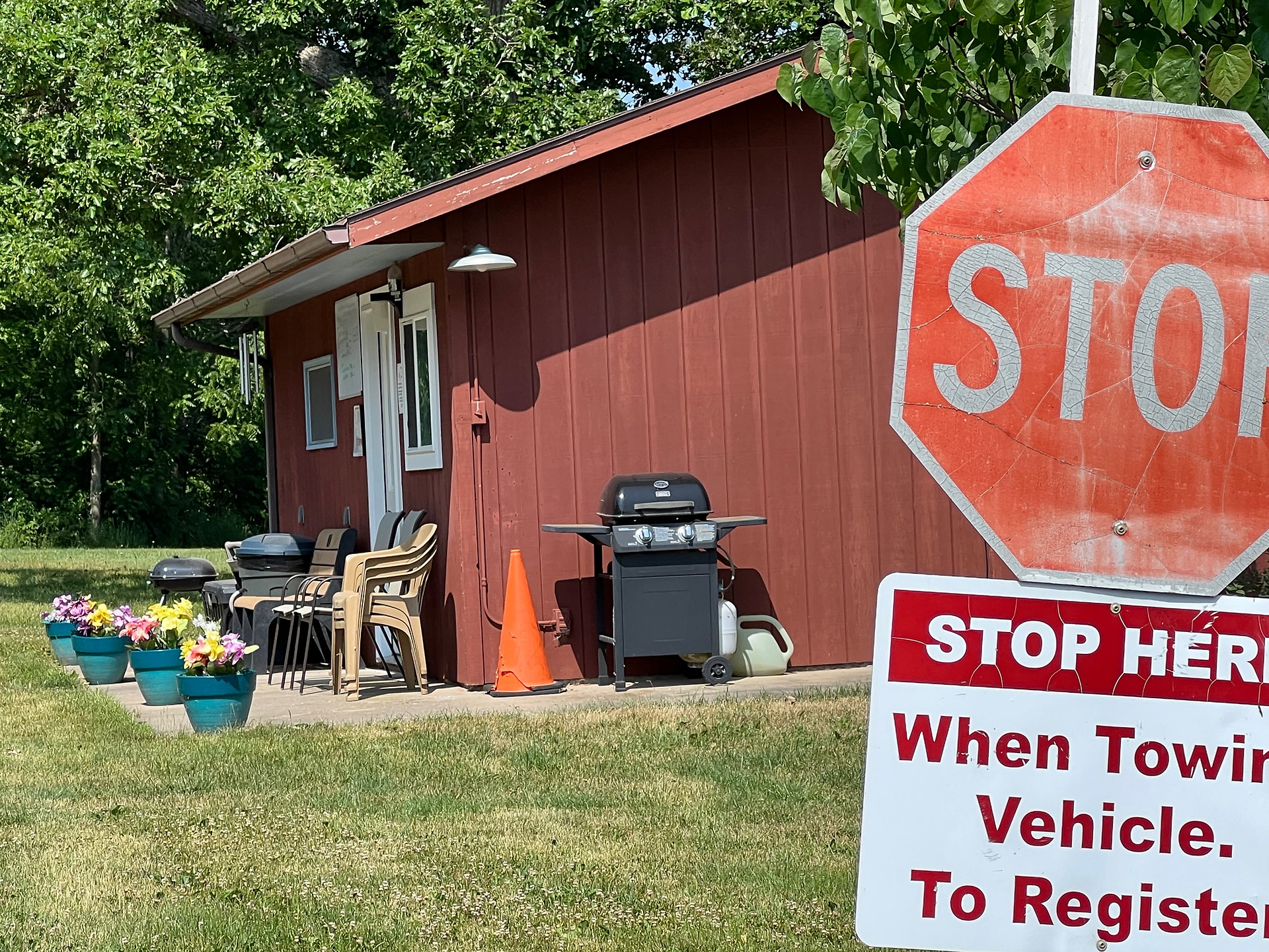 The image shows a small, red wooden building with a stop sign in the foreground. The stop sign is weathered and has a smaller sign beneath it that reads "STOP HERE When Towing Vehicle. To Register." The building has a white door and window, with a small overhang above the door. There are several plastic chairs stacked outside the building, along with a grill and some other outdoor equipment. To the left, there are a few potted plants with colorful flowers arranged along the side of the building. The area around the building is grassy and surrounded by lush green trees, creating a welcoming and well-maintained appearance.