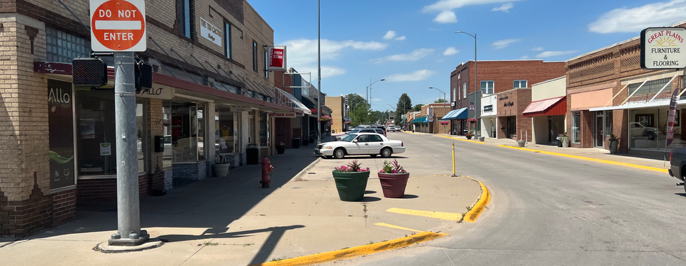 A small-town street lined with brick buildings housing various businesses, including a bookstore and a furniture store. The sidewalk is wide, and there are potted plants along the curb. A "Do Not Enter" sign is visible at the intersection. The sky is clear with a few scattered clouds, and the street is quiet with minimal traffic, creating a quaint and peaceful atmosphere.