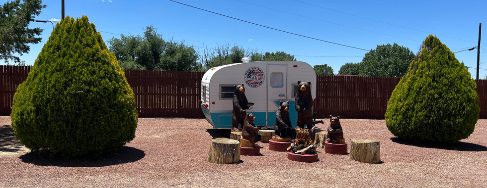 The image depicts a small vintage trailer parked on a gravel surface, decorated with a Route 66 sign on its side. In front of the trailer, there is a charming display of wooden bear carvings arranged around a faux campfire, creating a whimsical camp scene. The area is bordered by two large, well-trimmed evergreen bushes and a tall wooden fence in the background. The setting is outdoors under a clear blue sky, giving a sense of a sunny day.